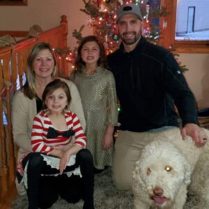 Jason Phillips with his family and their dog smiling in front of their Christmas tree.