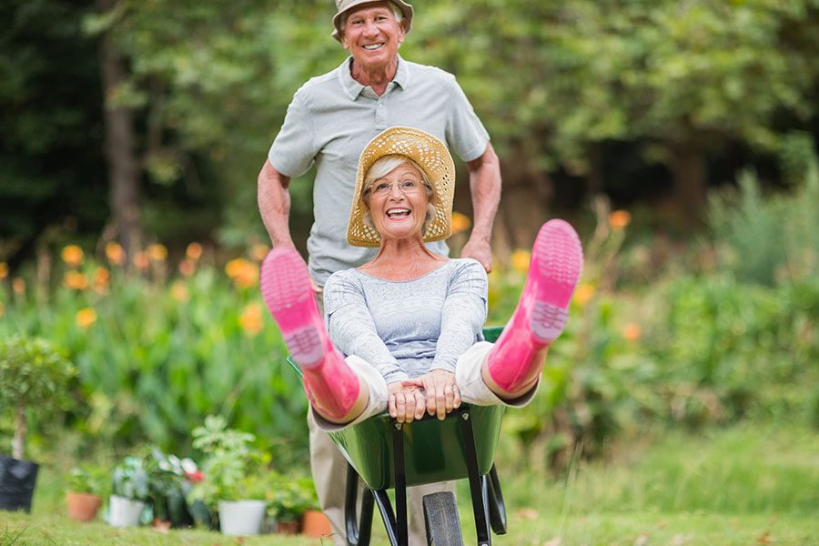 Contact - Portrait of a Joyful Mature Man Having Fun Pushing His Wife in a Wheelbarrow in the Garden