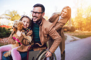 Dad in a wheelchair with his daughter and her puppy in his lap being pushed by his wife enjoying the security of their employee benefits insurance.