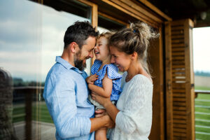 A young couple smiling while holding their daughter outside their home enjoying the peace of mind they get from their family insurance plan.