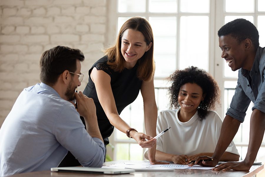 Defined Contribution - Midwest Select Insurance Group LLC - Woman Smiling and Explaining Benefits to Her Coworkers While They Standing and Sit at a Conference Table in the Office