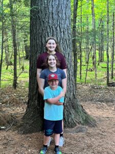 Jenna Deprey's three kids lined up smiling next to a white pine tree at their campground.