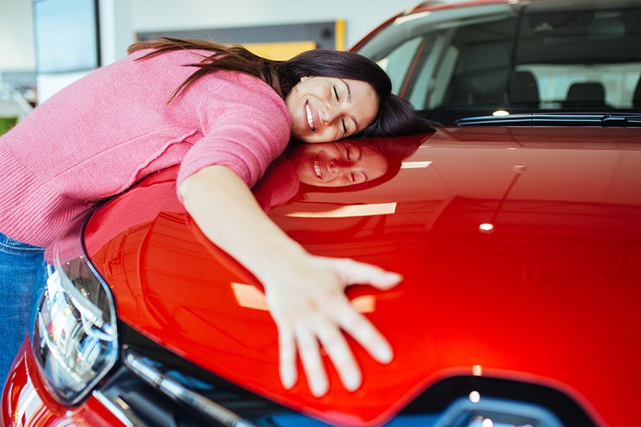 A happy woman smiling while hugging her new shiny red sedan.
