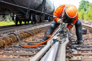 Interstate railroad worker welding train tracks together wearing an orange vest and black shin padding.