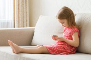 Young girl in a pink dress sitting on a couch using a tablet could be a victim of identity theft.
