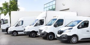 Five white business vans and trucks parked outside building are protected by commercial auto insurance.