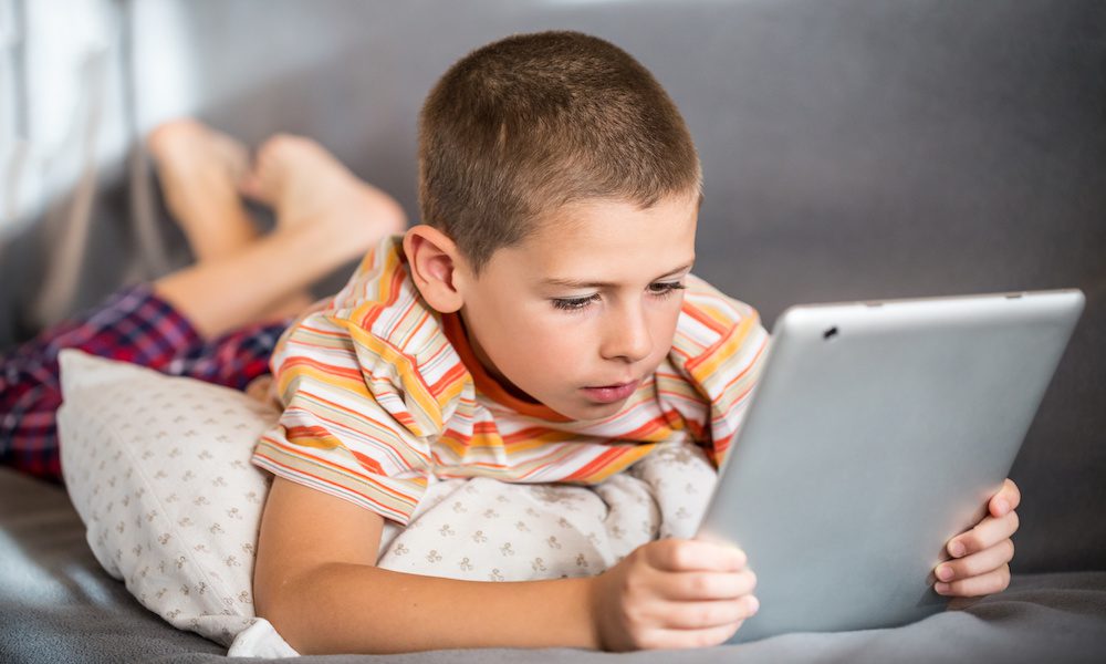 Boy in striped orange shirt playing on his tablet on a couch could be a victim of child identity theft.
